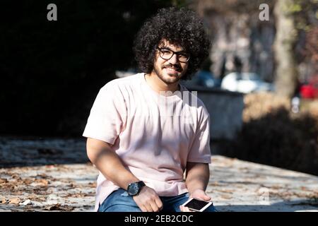 Porträt von schöner junger Afro Mann sprechen berühren Telefon oder Smartphone für den Anruf verwenden und im Herbst draußen sitzen Stockfoto