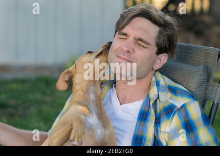 Hund küsst jungen Mann trägt ein kariertes Hemd aus gelb und blau in einem Hinterhof. Stockfoto