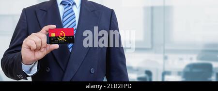 Zugeschnittenes Bild des Geschäftsmannes, der Plastikkreditkarte mit gedruckter Flagge von Angola hält. Hintergrund unscharf. Stockfoto