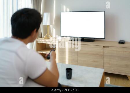 Ein asiatischer Mann hält eine FERNBEDIENUNG UND drückt den Sender, während er auf der Couch im Wohnzimmer zu Hause Fernsehen schaut. Stockfoto