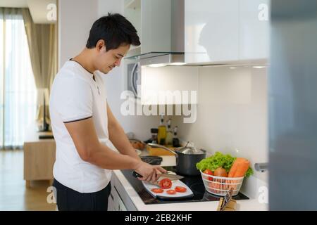 Ein asiatischer Mann schneidet Tomaten auf einer Küchentheke, um sich für das Abendessen zu Hause vorzubereiten. Stockfoto