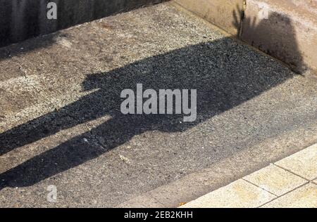Der Schatten einer Frau fällt auf die raue Oberfläche aus Asphalt und Beton, in harten und dunklen Farben Stockfoto