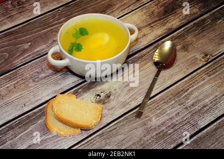 Tiefer runder Teller mit heißer Hühnerbrühe mit Ei und Petersilie auf einem Holztisch, Löffel und zwei Scheiben Weißbrot nebeneinander. Blick von oben. Stockfoto