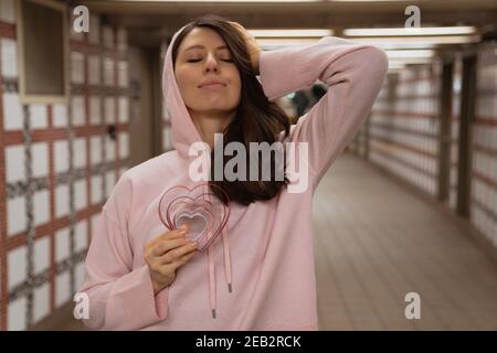 Junge schöne Frau mit geschlossenen Augen, hält Herzen in der Hand, trägt rosa Kapuzenpullover. Fröhliche, lustige Ausdrücke, Nahaufnahme Porträt. Valentinstag conce Stockfoto