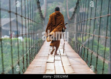 Ein Mönch mit einem Spazierstock Spaziergang auf einer hölzernen Hängebrücke in einem kleinen Dorf in Mae Hong Son, Thailand. Stockfoto