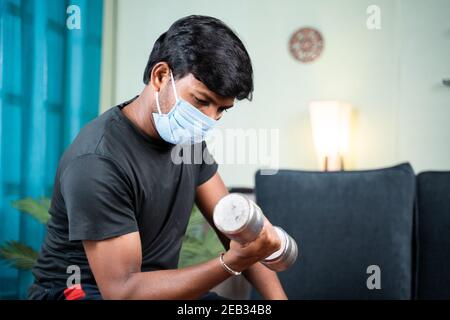 Junger Mann mit Gesichtsmaske beschäftigt in der Arbeit oder Doing Übung mit Hantel zu Hause - Konzept von zu Hause Fitness-Studio wegen Coronavirus covid-19 Pandemie Stockfoto