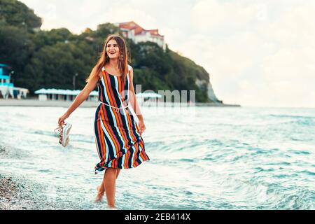 Sommerzeit. Eine glückliche Frau im Kleid geht am Strand entlang, hält ihre Sandalen in der Hand. Konzept der Sommerferien und Spaß. Stockfoto