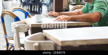 Ein Mann in einem grünen T-Shirt liest die Morgenzeitung und hält sie in den Händen. Eine weiße Kaffeetasse auf dem Tisch. Ruhe auf einem Café oder Restaurant Terrasse i Stockfoto