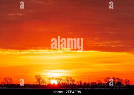 Sonnenaufgang, der durch ferne Hecken und Bäume am Horizont ganz unten im Rahmen erscheint. Himmel ist ein dramatisches Rot und Gelb mit einer dicken Wolkenschicht über der Sonne auch rot gefärbt. Stockfoto