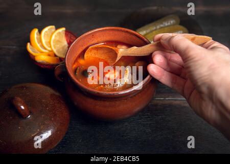 Frau schaufelt hausgemachte traditionelle russische Suppe Soljanka mit Fleisch, Würstchen, Gemüse, Gurken und Oliven mit Zitrone in keramischen Topf mit Holzlöffel Stockfoto