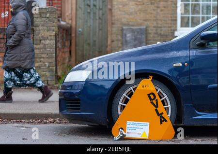 Southampton, Großbritannien, 11. Februar 2021. Ein Padestrian geht an einem geparkten blauen Auto mit einer gelben DVLA-Autoladerklammer vorbei, die am Beifahrer-Seitenrad in einer Wohnstraße in Southampton, Hampshire, befestigt ist. Stockfoto