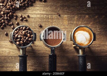Kaffee in seinen verschiedenen Formen von rohen Bohnen über gemahlenen Kaffee bis hin zu Cappuccino in drei Portafiltern auf rustikalem Holzhintergrund. Stockfoto
