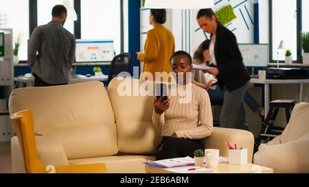 Müde schwarze Frau im Gespräch mit Kollegen mit Videoanruf halten Smartphone mit Wiraless-Headset sitzen auf der Couch im Büro. Vielfältiges Team plant neues Finanzprojekt in modernen Unternehmen Stockfoto