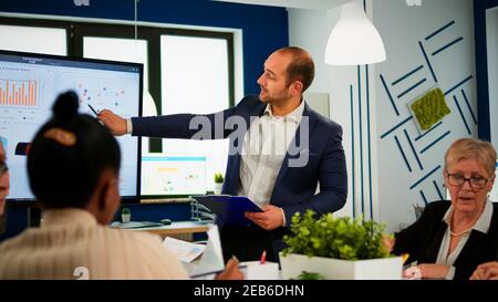 Comapany CEO hält Zwischenablage und zeigt auf digitale interaktive Whiteboard mit Wachstumsanalyse Statistiken und Daten. Verschiedene Personen, die während der Konferenz im Broadroom arbeiten und Startprobleme diskutieren Stockfoto