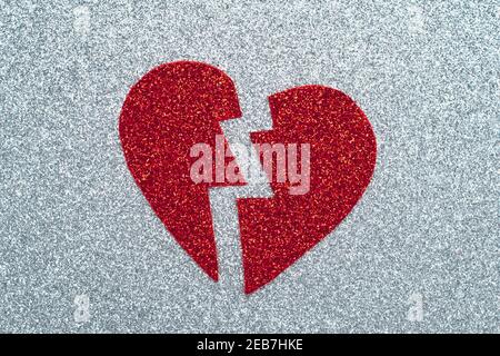 Gebrochenes rotes Herz auf grauem Glitter-Papier, ein Zerbrechungskonzept. Lametta-Applikation. Ein Symbol der Liebe, Valentinstag, romantische Gefühle und Emotionen. Abstra Stockfoto