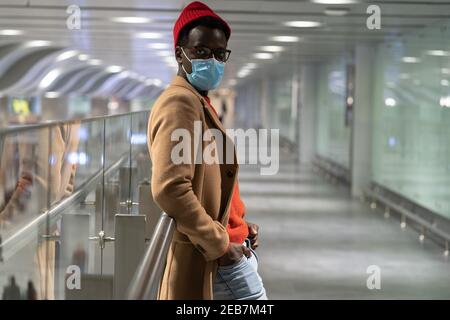Trendy afrikanischen Hipster Mann Blick auf Kamera, im Flughafen-Terminal stehen, tragen Gesichtsmaske. Covid-19 Stockfoto