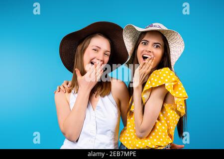 Portrait von überraschten Freundinnen im Sommer helle Kleidung und Hüte Blick auf die Kamera auf einem blauen Hintergrund. Stockfoto