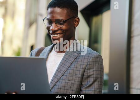 Fröhlicher afroamerikanischer Mann, der lächelt und im Stehen einen Laptop benutzt Im Freien Stockfoto