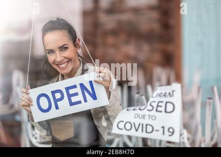 Fröhliche Kellnerin lächelt über dem offenen Schild, als das Restaurant nach einer Corona-Pandemie wieder öffnet. Stockfoto