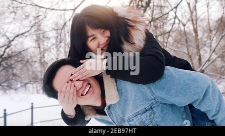 Junger Mann gibt Huckepack Fahrt zu seiner Freundin im Park an verschneiten Wintertag. Newlywed asiatisch pärchen mit Spaß draußen. Hochwertige Fotos Stockfoto