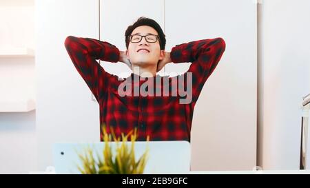 Junge positive asiatische Kerl lehnte sich zurück in seinem Stuhl nach Beendigung der Arbeit an seinem Laptop zu Hause. . Hochwertige Fotos Stockfoto