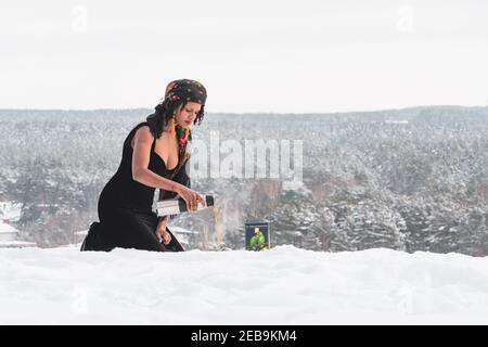 Schönes Mädchen mit schwarzem Kleid trinken heißen Kaffee oder Tee Auf dem Schnee im Winter mit Waldbäumen bedeckt Schnee und gefrorener Fluss im Hintergrund Stockfoto