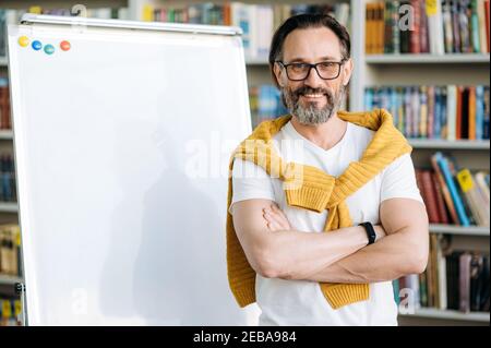 Portrait von fröhlichen Freiberufler in stilvoller Kleidung am Arbeitsplatz. Glücklicher Geschäftsmann in Brillen, der in der Nähe von Markiertafel im Büro steht, direkt auf die Kamera schaut und lächelt Stockfoto