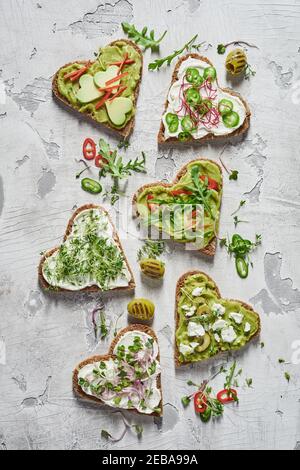 Verschiedene herzförmige Tapas, Canapes oder Cichitti mit Avocadobirne, frischen Kräutern, Käseaufstrich und Chili in einer flachen Lay-Down-Ansicht auf einem Peeling Whi Stockfoto