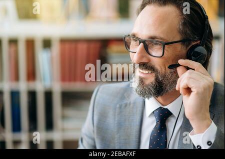 Nahaufnahme Porträt von zufriedenen hilfsbereiten reifen männlichen Callcenter-Operator, Mitarbeiter des Support-Centers in formellem Anzug und Headset, sitzen im Büro freundlich lächelnd. Arbeit am Support-Service Stockfoto