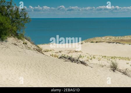 Sanddünen mit Grassträuchern im Sonnenlicht am Meer türkisfarbenes Meer und Himmel mit Wolken. Sommerliche Landschaft mit hellen Sanddünen und Meer. Blaues Meer und Stockfoto