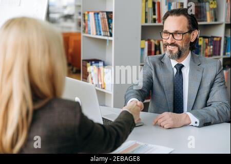Freudig reifen Mann schüttelt Hände mit blonden Haaren Frau nach erfolgreichen Verhandlungen. Schöner Mann in stilvollen formellen Anzug auf Interview im modernen Büro, lächelnd, glücklich über einen neuen Job zu bekommen Stockfoto