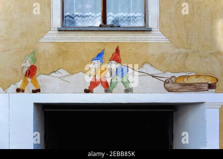 Gnome Baking … Vor dem Hintergrund der Berggipfel werden die frischen Brote aus den Brotbacköfen der kleinen Leute von Märchengnomen in bunten spitzen Hüten auf einem Schlitten über die Sgraffito-Front eines der alten Häuser in Guarda gezogen, Ein malerisches, überwiegend rätoromanisch sprechendes Dorf im Unterengadintal im Kanton Graubünden, Ostschweiz. Häuser hier stammen aus dem frühen 1600s, mit vielen mit Gemälden und Sgraffito Kunst auf ihrer façades. Das Dorf hat eine Schweizer nationale Auszeichnung für seinen Erfolg bei der Erhaltung seines reichen architektonischen und kulturellen Erbes gewonnen. Stockfoto