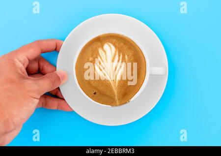 Draufsicht Hand Mann hält weißen Kaffeeuntersetzer mit Cappuccino-Kaffee und cremige Form in weißer Tasse. Der Handmann hält die Kaffeetasse auf dem blauen Tisch im Kaffee Stockfoto