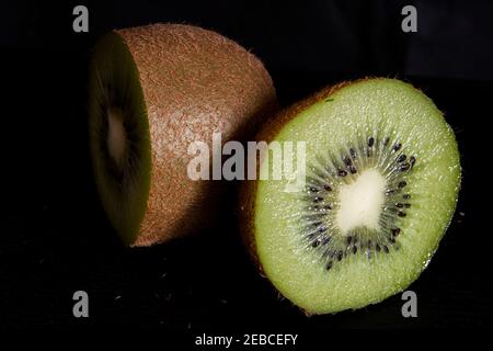 Kiwi-Frucht in zwei Hälften geschnitten auf schwarzem Hintergrund, mit sichtbarem weißen Kern und schwarzen Samen. Die Frucht ist nicht geschält, die braune Haarschale ist sichtbar Stockfoto
