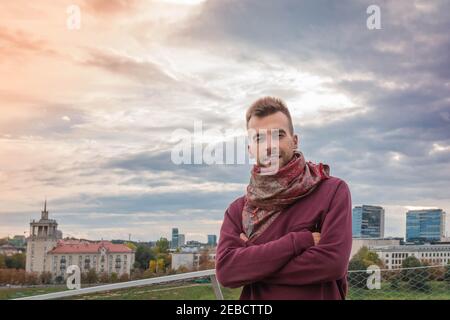 Porträt eines gutaussehenden Mannes mit gekreuzten Armen am dramatischen Himmel und städtischen Hintergrund. Attraktiver Mann, der auf der Terrasse am bunten Himmel und im Hintergrund der Stadt steht. Stockfoto