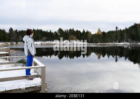 Frau, die auf einer verschneiten Holzbrücke steht und auf eine gefrorene schaut see Stockfoto