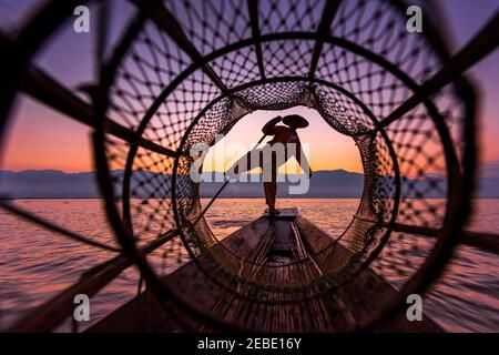 Lake Inle Fischer in traditioneller Kleidung und Hut balanciert ein Bein während des Fischens, geschossen durch Netz während lebhafter Sonnenaufgang außerhalb Yangon, Myanmar Stockfoto