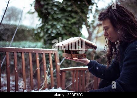 Asiatische Person im Schnee hält hölzerne Vogelhaus hinzuzufügen Samen im Hinterhof Stockfoto