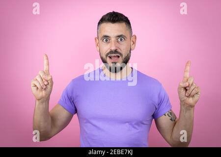 Junger schöner Mann trägt lässiges T-Shirt über rosa Hintergrund erstaunt Und überrascht, die Kamera zu betrachten und mit nach oben zu zeigen Finger und angehobene Arme Stockfoto