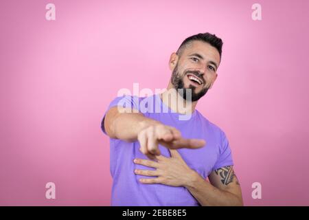 Junger, hübscher Mann mit lässigem T-Shirt auf rosa Hintergrund lachend auf Sie, zeigt den Finger auf die Kamera mit der Hand über den Körper, Scham Ausdruck Stockfoto