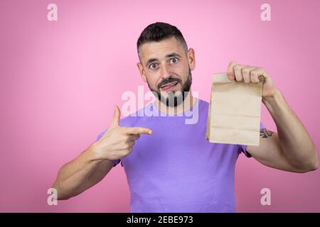 Junger hübscher Mann trägt lässiges T-Shirt über rosa Hintergrund lächelnd, überrascht und zeigt Papiertüte in ihrer Hand Stockfoto