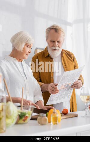 Ältere paar lesen Nachrichten beim Kochen auf verschwommenem Vordergrund in Küche Stockfoto