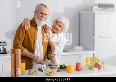 Lächelnde Frau umarmt älteren Mann schneiden Gemüse in der Nähe von Orangensaft Und Schüssel in der Küche Stockfoto