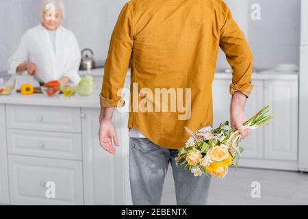 Älterer Mann versteckt Blumen in der Nähe Frau Kochen in der Küche auf Unscharfer Hintergrund Stockfoto