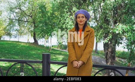 Glückliche Frau in stilvoller Baskenmütze und Mantel in der Nähe von Zaun stehen Im Park Stockfoto