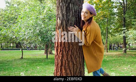 Fröhliche trendige Frau lächelt an der Kamera, während umarmt Baumstamm Im Park Stockfoto