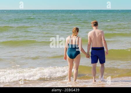 Paar machen sich bereit für das Schwimmen im winkenden Meer. Frau und Mann halten ihre Hände zusammen gehen, um in Welle spritzt Meer schwimmen. Pairing geht weiter int Stockfoto