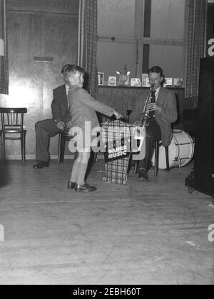 1950s, historisch, in einem Veranstaltungsraum bei einer Geburtstagsparty zeigt ein kleiner Junge mit Jacke, Shorts und langen Socken auf ein Schild "Dance Music", wo ein Mann neben einem Klavier sitzt und Trompete spielt, England, Großbritannien. Zu dieser Zeit war 'Big Band' Musik die populäre und tragende Säule der Musik und dann kam Skiffle, was zu Rock and Roll führte. Stockfoto