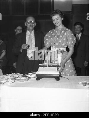 1950s, historisch, neben ihrem stolzen Großvater stehend, eine elegante junge Frau, die ein stilvolles gepunktetes oder gepunktetes Kleid trägt, das ihren Geburtstagskuchen auf ihrer Geburtstagsparty in einem Veranstaltungsraum eines Hotels, England, Großbritannien, schneidet. Stockfoto