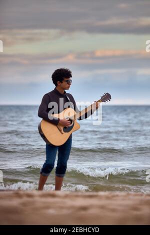 Junge kaukasische Teenager spielen akustische Gitarre am Strand. Sonnenbrillen und dunkle Kleidung. Benetzung ihrer Hose mit einer Welle Stockfoto